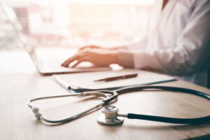 Close up of a stethoscope on a desk with a doctor typing on a laptop in the background